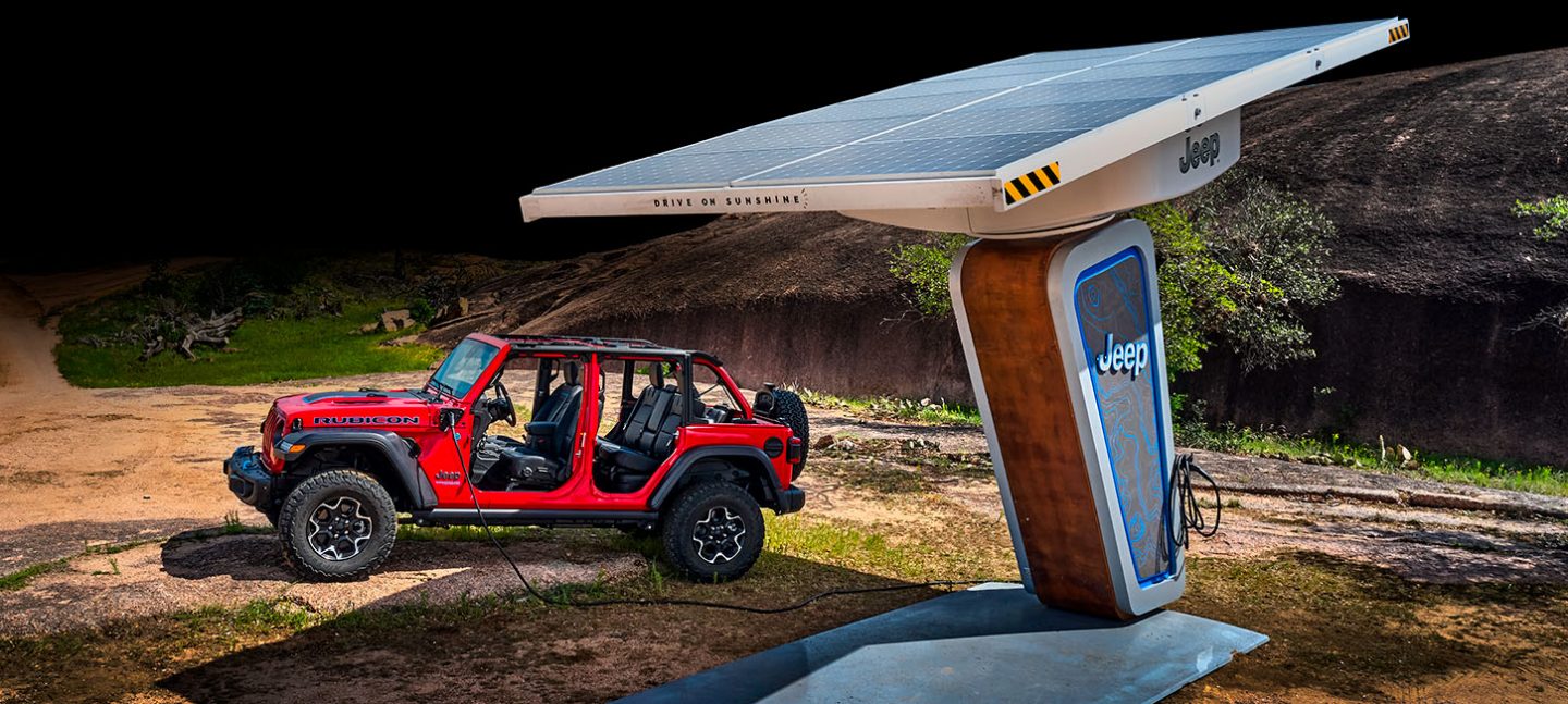 A 2022 Jeep Wrangler Rubicon 4xe parked next to a Jeep Brand solar charging station in the woods as it charges its electric motor battery.